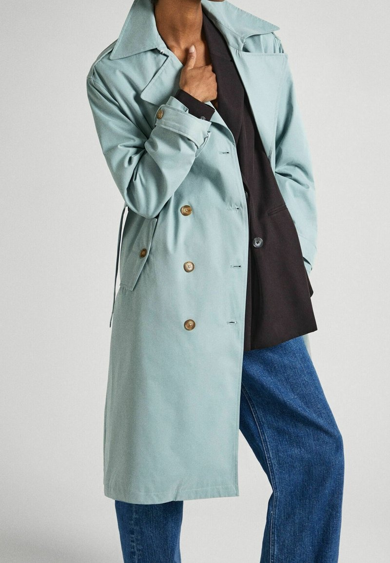 Personne portant un trench bleu clair avec des boutons beige sur un blazer noir et un jean bleu, posant avec une main près du col.