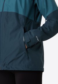 Waterproof jacket in dark teal with a lighter top section. Features zippers, pockets, and "DRY VENT" label on the cuff. Smooth texture.