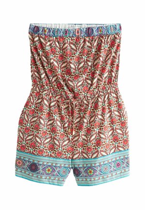 Kobieta z kręconymi włosami ubrana w kwiatowy kombinezon bez ramiączek z wzorzystymi obszyciami, stojąca z rękami na biodrach na beżowym tle.