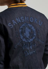 Veste en denim foncé avec col à rayures jaunes, design bleu brodé avec le texte "SANSHOKU", motif de poisson koi et détails "RISING GRACE".
