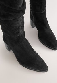 Schwarze kniehohe Wildlederstiefel mit spitzen Zehen, Blockabsätzen und seitlichen Reißverschlüssen vor hellem Hintergrund.