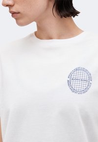 Άτομο που φοράει ένα λευκό μπλουζάκι με μπλε κυκλικό λογότυπο που γράφει "Monaco Racquet and Health Club" και "Björn Borg" στο στήθος.