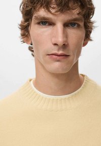 Jasnożółty sweter z prążkowanym, okrągłym dekoltem. Gładka dzianina. Modelka o potarganych brązowych włosach i niebieskich oczach. Zbliżenie.