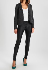 Černý texturovaný blazer s jedním knoflíkem, v kombinaci s lesklými černými legínami s ribbovanými detaily. Modelka má na sobě tyrkysové vysoké podpatky.