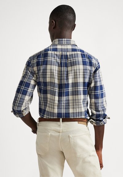 Μπλε και κρεμ πουκάμισο με καρό σχέδιο, γιακά με κουμπιά, μανίκια τυλιγμένα και ευθεία οπίσθια άκρη. Συνδυασμένο με ανοιχτόχρωμο παντελόνι.
