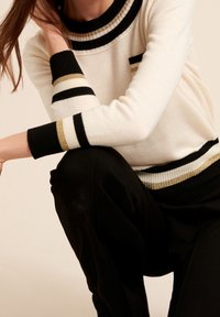 Maglione bianco con accenti a strisce nere e oro nel collo e nei polsini; texture a coste; abbinato a pantaloni neri.
