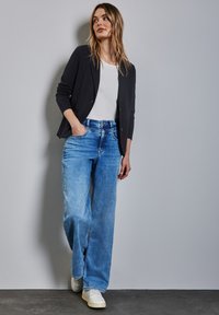 Cardigan nero sopra una maglietta bianca, abbinato a jeans a gamba larga azzurri e sneaker bianche. Texture liscia; vestibilità casual.