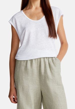 Kobieta w białej bluzce z krótkim rękawem i dekoltem w serek, włożonej do wysokich, luźnych szarych spodni w kratę, z rękami luźno opuszczonymi.