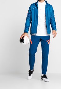 Nike urheiluvaatteet, joissa on sininen takki, valkoinen paita, siniset housut oransseilla yksityiskohdilla ja musta-valkoinen jalkapallo.