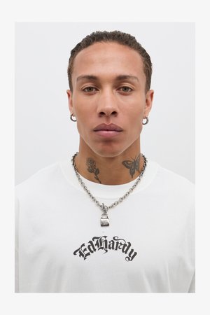 T-shirt blanc avec logo "Ed Hardy" en noir, collier en chaîne argenté et pendentif en forme de cadenas. Présente des tatouages d'une rose et d'un papillon sur le cou.