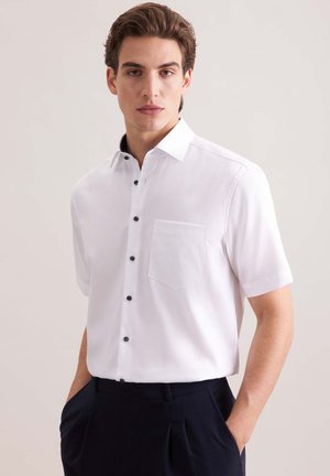 Jeune homme portant une chemise blanche à manches courtes avec des boutons noirs et un pantalon bleu marine, debout devant un fond clair uni.