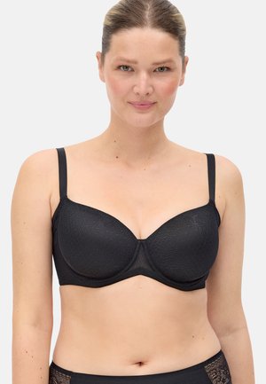 Mujer con el cabello recogido, vistiendo un sujetador negro con aro, copas ligeramente texturizadas y ropa interior negra con detalles de encaje, sobre un fondo blanco.