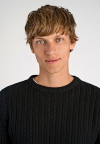 Zwarte gebreide trui met een textuurpatroon, ronde hals en geribbelde manchetten. Het model heeft lichtbruin haar en blauwe ogen.