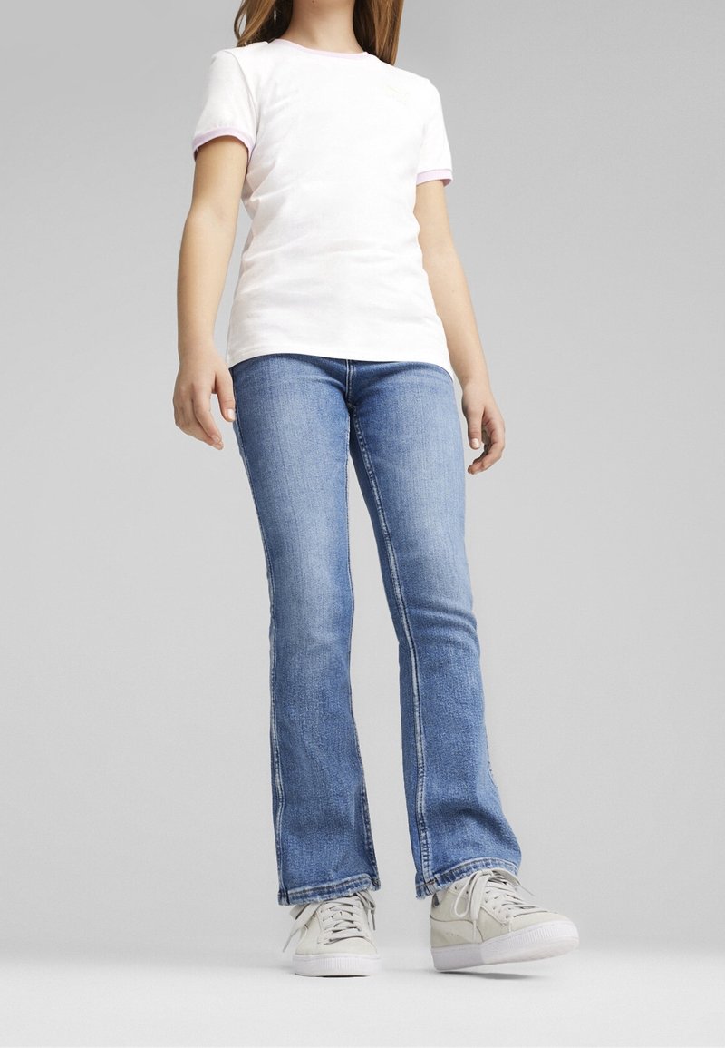 Personne portant un t-shirt blanc, un jean bleu légèrement évasé et des baskets blanches, debout devant un fond gris uni.
