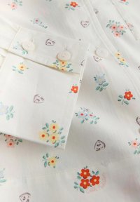 Biely textil s malými červenými, žltými a modrými kvetinovými vzormi a roztrúsenými malými motívmi srdiečok, s bielymi gombíkmi na manžete košele.