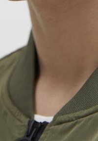 Khaki kurtka bomberka z prążkowanym kołnierzem, zamkiem błyskawicznym i teksturowaną tkaniną widoczną w okolicy szyi i ramion.