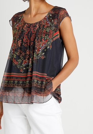 Vrouw draagt een mouwloze zwarte doorschijnende blouse met rode en groene bloemenpatronen, gecombineerd met een witte broek, hand in de zak.