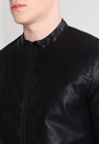 Giacca di pelle nera con colletto alto, dotata di una cerniera frontale e una texture liscia. Il design è aderente, mostrando uno stile minimalista.