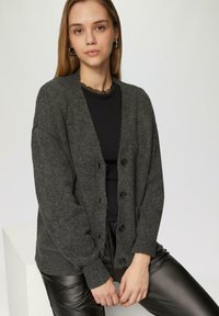 Dunkelgrauer, gestrickter Cardigan mit V-Ausschnitt, Knopfverschluss und gerippten Bündchen. Über einem schwarzen Oberteil getragen, kombiniert mit schwarzen Lederhosen.