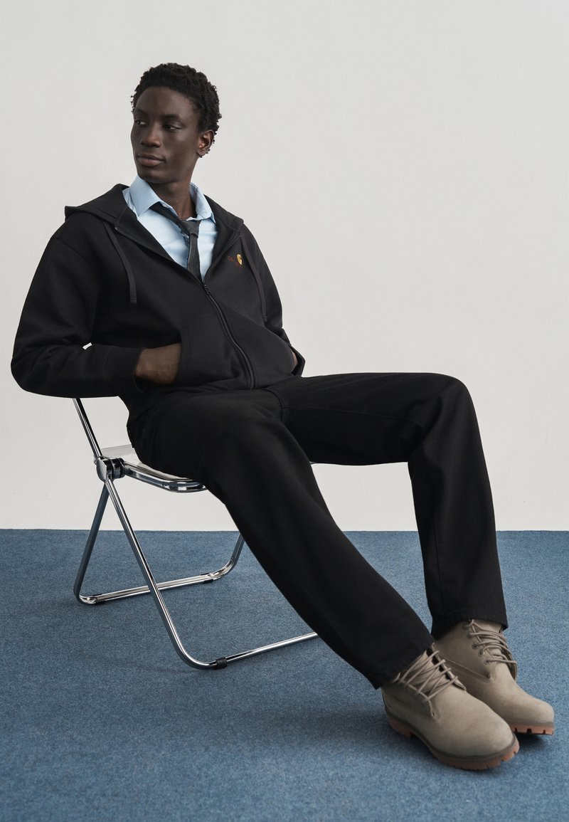 Άνδρας με μαύρη φούτερ με κουκούλα, ανοιχτό μπλε πουκάμισο, μαύρη γραβάτα, μαύρο παντελόνι και μπεζ μπότες, που κάθεται σε μεταλλική αναδιπλούμενη καρέκλα σε μπλε χαλί.