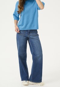Femme portant un pull à manches courtes bleu clair, un jean à jambes larges bleu et des baskets blanches, debout devant un fond uni.