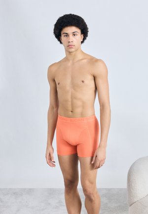 Svetlo oranžové boxerky s hladkou textúrou a elastickým pásom, na ktorom je nenápadné logo "BJÖRN BORG". Model je zobrazený z profilu.