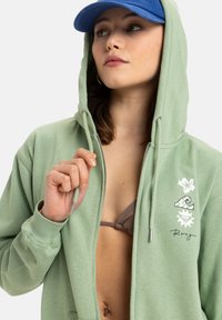 Ljusgrön zip-up hoodie med dragsko på huvan, blommiga och vågmönstertryck, samt en avslappnad passform. Bärs över en brun bikinitopp.