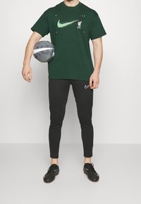 Zielona koszulka Nike z dużym logo, grafika Liverpoolu; czarne spodnie dresowe Nike; trzymający szary piłkę nożną; czarne obuwie z pomarańczowymi podeszwami.
