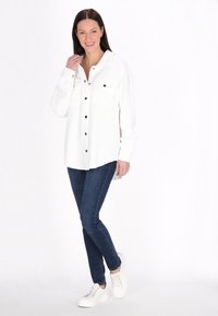 Camicia bianca con tessuto a trama, quattro bottoni neri sul davanti, due tasche sul petto, abbinata a jeans blu e sneaker bianche.