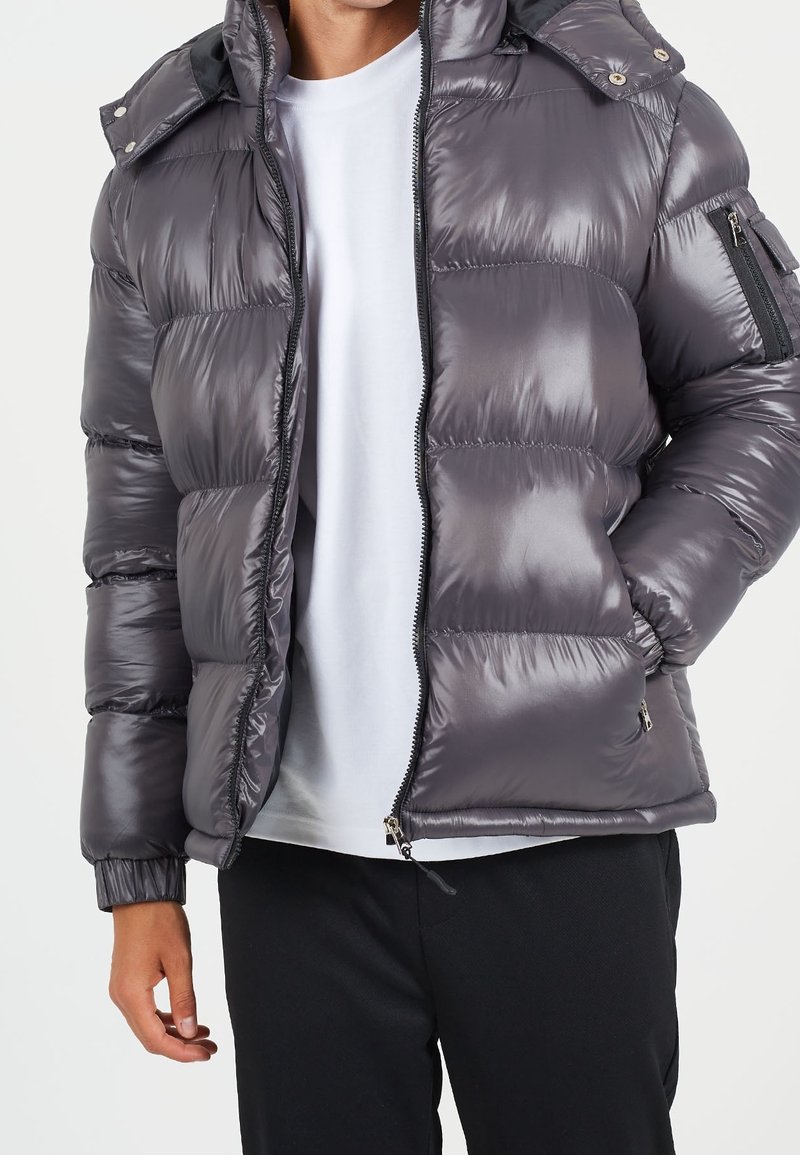 Metallisk grå vadderad jacka med huva, dragkedja framtill och sidofickor. Designad med elastiska manschetter och en slät, glänsande yta.