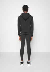Zwarte windjack met capuchon, slim-fit ontwerp. Gedragen over zwarte leggings en sportieve sneakers. Gladde textuur, minimalistische stijl.