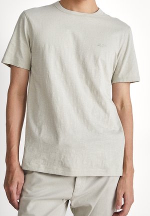 Beiges T-Shirt mit kurzen Ärmeln und Rundhalsausschnitt, getragen von einer Person, kombiniert mit passenden beigen Hosen, schlichtes Design mit dezentem Brustlogo.