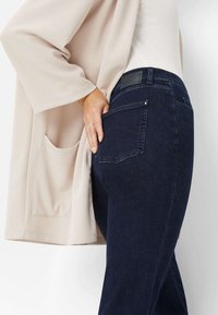 Jeans blu scuro con una texture liscia e un delicato dettaglio di cucitura, abbinati a un cardigan beige chiaro con una tasca frontale.