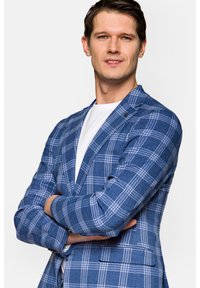 Niebieski blazer w kratę o dopasowanym kroju, z jednym guzikiem, klapami i dwiema przednimi kieszeniami. Nosi się go na białej koszuli.