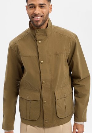 Mężczyzna uśmiechający się, ubrany w brązową zapinaną na guziki kurtkę z klapkami na kieszeniach, stojący na tle jednolitego białego tła.