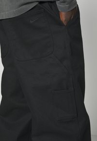 Black cargo pants made of durable fabric, featuring side and rear pockets, double stitching, and a subtle Nike logo on the back pocket.