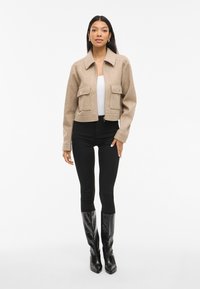 Giacca beige cropped con colletto e due tasche frontali, abbinata a jeans skinny neri e stivali neri al ginocchio. Texture liscia.