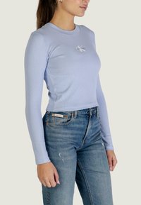 Top azzurro chiaro a maniche lunghe, corto, in tessuto a costine con un piccolo logo. Abbinato a jeans blu a vita alta.