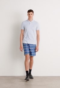 Ανοιχτό μπλε κοντομάνικο πόλο μπλουζάκι, μπλε σορτς με καρό σχέδιο, μαύρες κάλτσες και μαύρα παπούτσια σε ανδρική φιγούρα σε απλό φόντο.