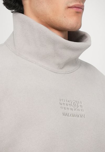 Světle šedý fleecový pulovr s vysokým límcem. Obsahuje embosovaná čísla a logo značky. Měkká textura, minimální detaily šití.