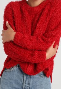Pull en tricot rouge flou avec des manches longues, un col rond et des fentes sur les côtés. Associé à un jean en denim bleu clair taille haute. Texture douce.
