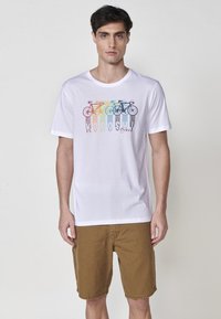 T-shirt bianca in cotone con una grafica colorata di una bicicletta. La maglietta ha maniche corte e una vestibilità rilassata. Il modello indossa pantaloni corti marroni.
