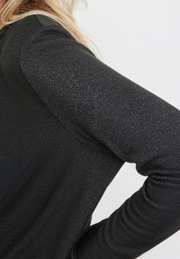 Svart långärmad topp med en texturerad tyg och en subtil lyster samt en figurnära design. Ärmen visar en smidig övergång från halslinningen.