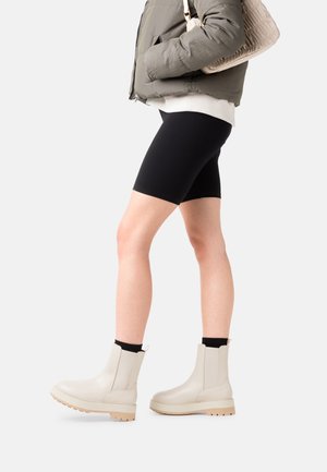 Persoon in beige enkellaarzen, zwarte sokken, zwarte shorts, een wit sweatshirt en een groene jas, met een beige handtas op de schouder.