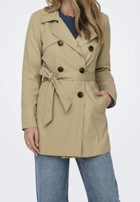 Trench beige avec taille ceinturée, fermeture à double boutonnage, col à revers, poches latérales et tissu texturé.