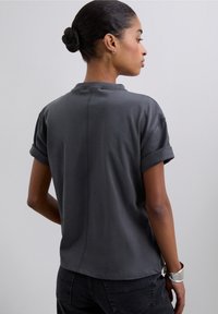 Tricou gri cu mâneci scurte, manșete rulotate, croială lejeră, detaliu cusătură pe spate și textură moale. Purtați împreună cu blugi închiși la culoare și brățară argintie.