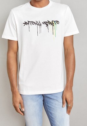 Personne portant un t-shirt blanc à manches courtes avec le texte en style graffiti noir "ANTONY MORATO" dégoulinant de peinture jaune et verte, associé à un jean bleu clair.