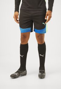 Schwarze Sportshorts mit blauem gemustertem Saum, kombiniert mit schwarzen Kniestrümpfen mit einem weißen Logo und schwarzen Fußballschuhen.