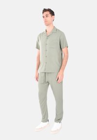 Camicia verde oliva chiaro a maniche corte con tasca, abbinata a pantaloni jogger coordinati. Entrambi realizzati in tessuto liscio; indossati con sneakers bianche.
