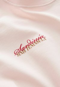 Camiseta de algodón rosa claro con texto bordado en rojo y dorado: "Sandinia" y "PENÍNSULA DE SINIS". Textura suave y corte clásico.